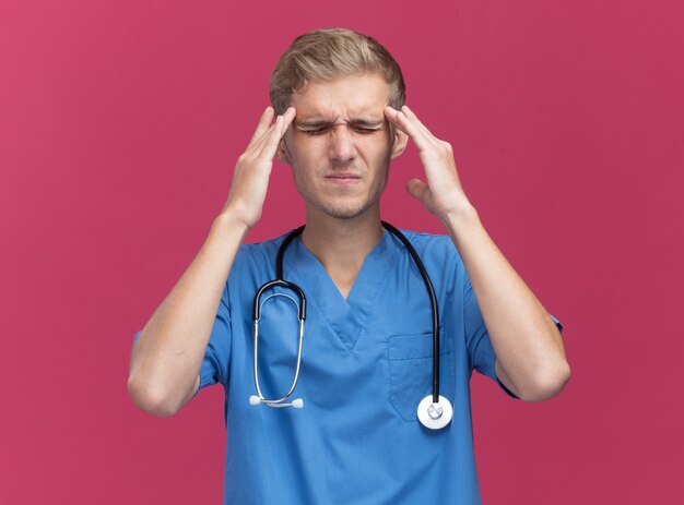 Unpleased with closed eyes young male doctor wearing doctor uniform with stethoscope putting fingers on aching temples isolated on pink wall