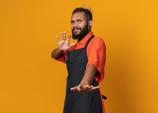 Free photo unpleased young afro-american male barber wearing uniform standing in profile view  doing refusal gesture isolated on orange wall with copy space