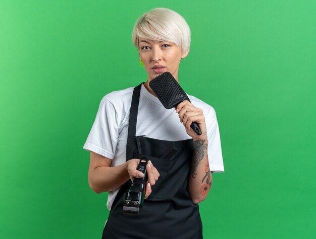 Unpleased young beautiful female barber in uniform holding hair clipper with comb isolated on green background