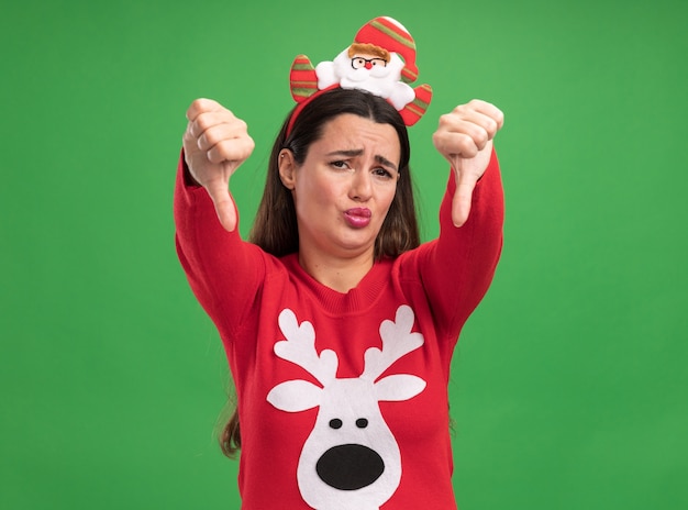 Free photo unpleased young beautiful girl wearing christmas sweater with christmas hair hoop showing thumbs down isolated on green background