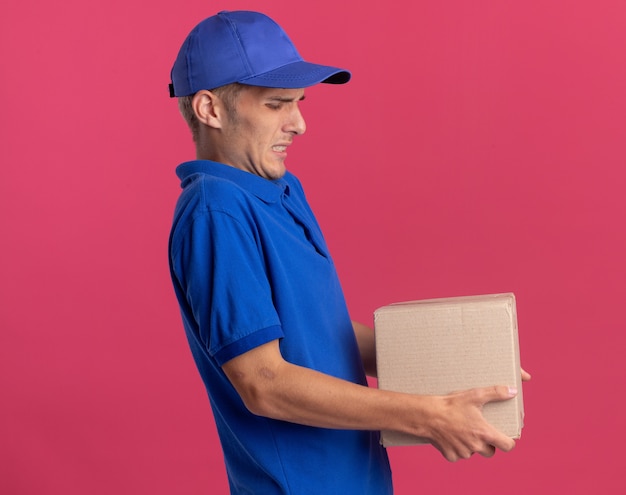 Unpleased young blonde delivery boy stands sideways holding heavy cardbox