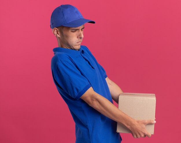 Unpleased young blonde delivery boy stands sideways holds heavy cardbox isolated on pink wall with copy space