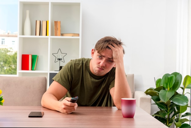 Free photo unpleased young blonde handsome man sits at table with phone and cup putting hand on forehead holding tv remote