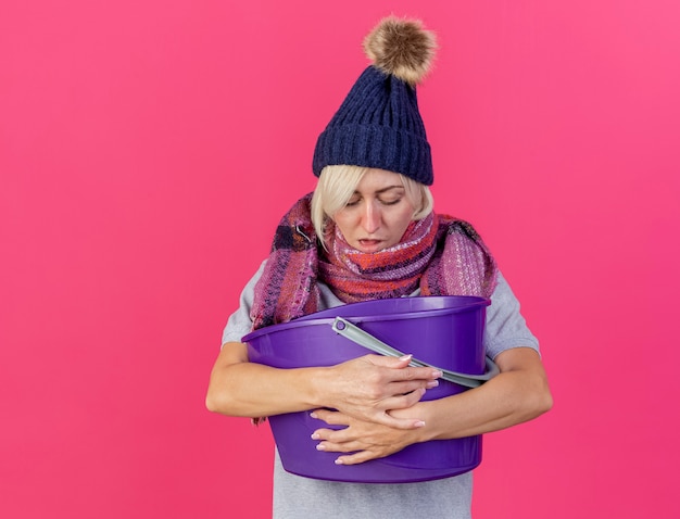 Free Photo unpleased young blonde ill slavic woman wearing winter hat and scarf holds and looks at plastic bucket having nausea isolated on pink wall with copy space