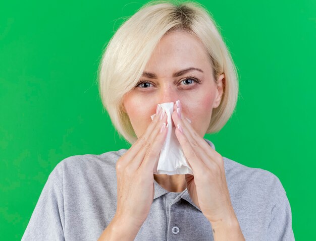 Unpleased young blonde ill slavic woman wipes nose with tissue isolated on green wall with copy space