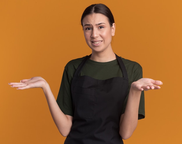 Unpleased young brunette barber girl in uniform holds hands open isolated on orange wall with copy space