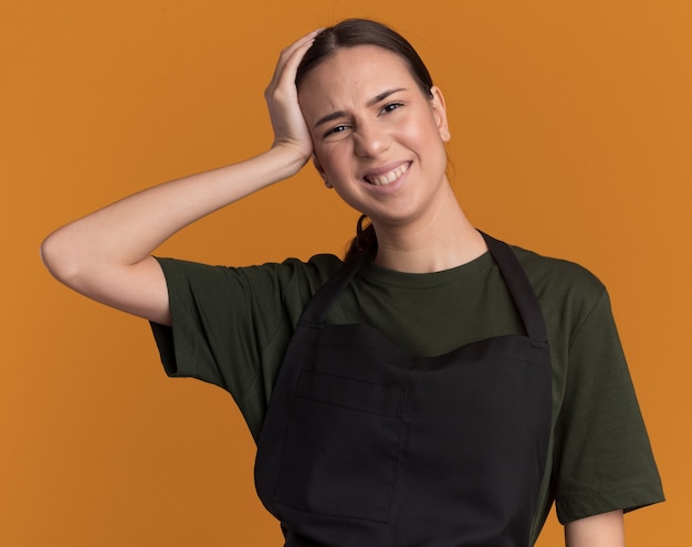 Unpleased young brunette barber girl in uniform puts hand on head and looks at camera on orange