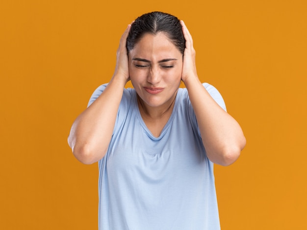 Unpleased young brunette caucasian girl covers ears with hands isolated on orange wall with copy space