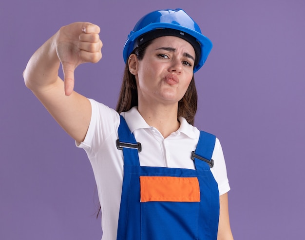 Unpleased young builder woman in uniform showing thumb down isolated on purple wall