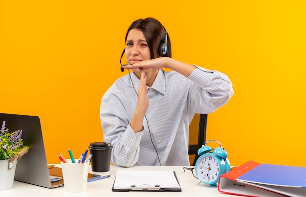 Free photo unpleased young call center girl wearing headset sitting at desk with work tools doing timeout gesture isolated on orange background