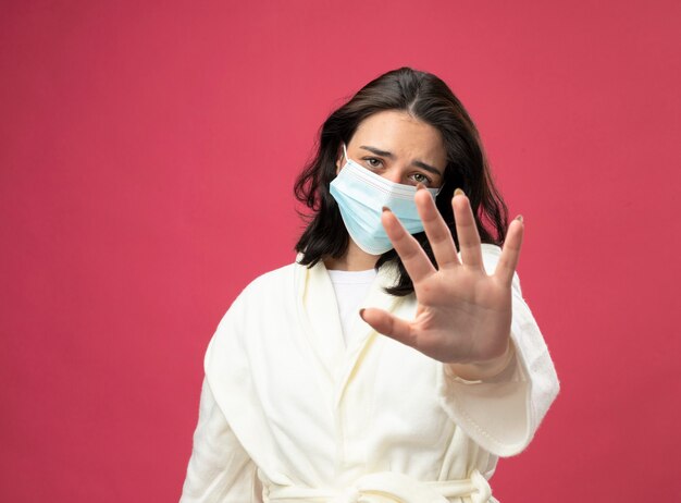 Unpleased young caucasian ill girl wearing robe and mask looking at camera stretching out hand towards camera gesturing no isolated on crimson background with copy space