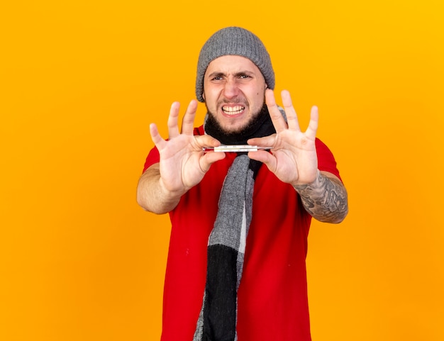 Free photo unpleased young caucasian ill man wearing winter hat and scarf holds thermometer isolated on orange wall with copy space