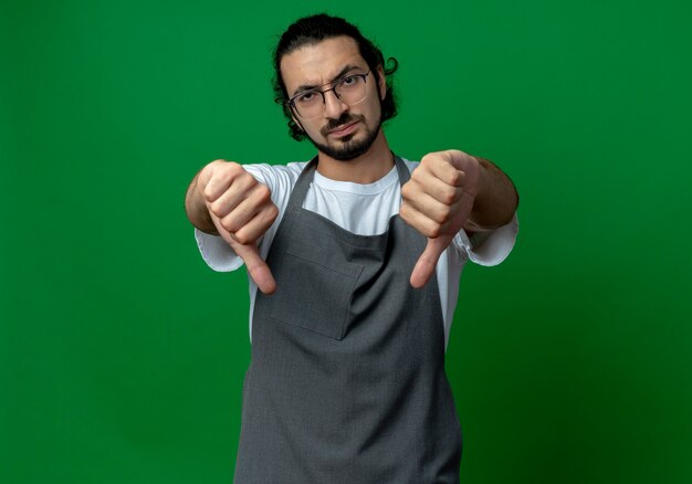 Unpleased young caucasian male barber wearing uniform and glasses pointing down isolated on green background with copy space