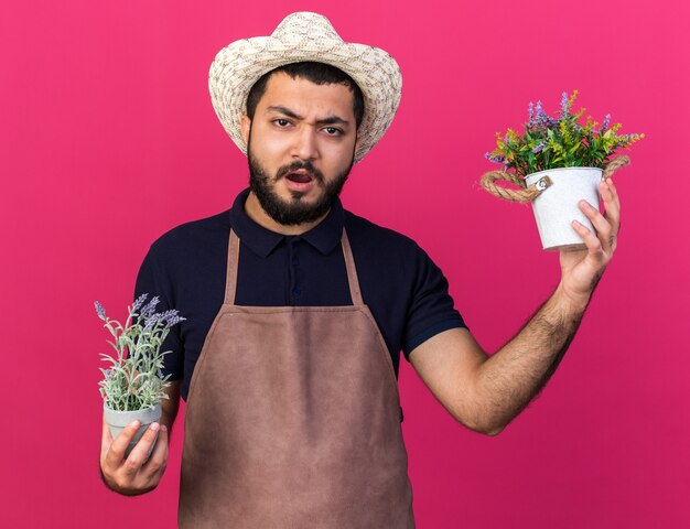 unpleased young caucasian male gardener wearing gardening hat holding flowerpots isolated on pink wall with copy space