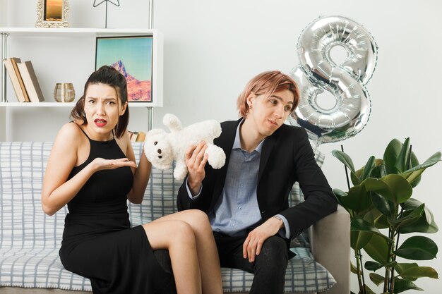 Unpleased young couple on happy women day with teddy bear sitting on sofa in living room