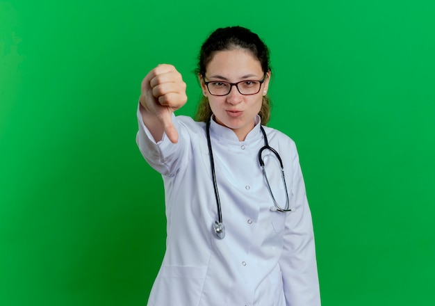 Unpleased young female doctor wearing medical robe and stethoscope and glasses  showing thumb down isolated on green wall with copy space