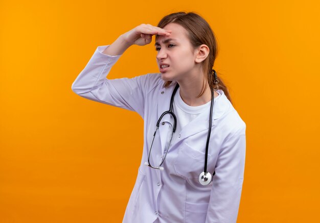 Unpleased young female doctor wearing medical robe and stethoscope putting hand near forehead and looking at distance on isolated orange wall