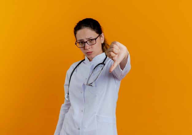 Unpleased young female doctor wearing medical robe and stethoscope with glasses showing thumb down isolated