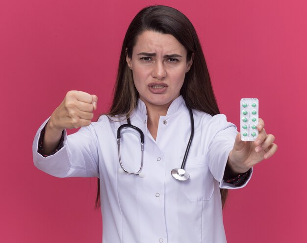 Unpleased young female doctor wearing medical robe with stethoscope holds pack of medicine