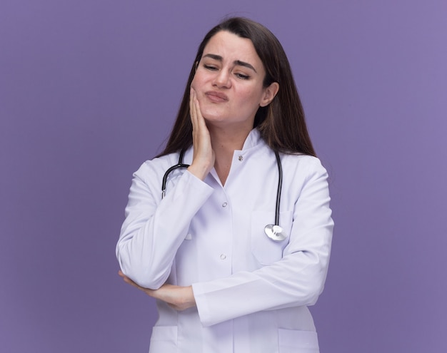 Free photo unpleased young female doctor wearing medical robe with stethoscope puts hand on chin and looks down isolated on purple wall with copy space