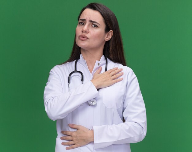 Unpleased young female doctor wearing medical robe with stethoscope puts hand on shoulder isolated on green wall with copy space