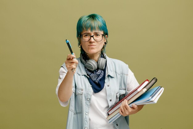 Unpleased young female student wearing glasses bandana and headphones around neck holding note pads looking at camera pointing at camera with pen isolated on olive green background