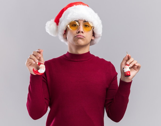 Free Photo unpleased young guy wearing christmas hat holding christmas toys isolated on white wall