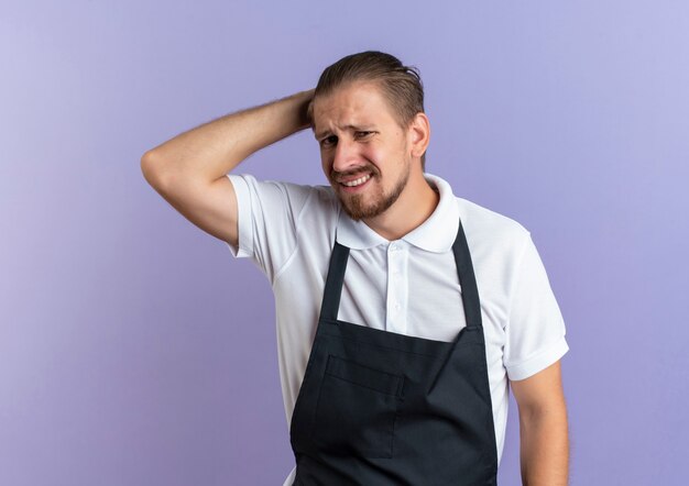 Unpleased young handsome barber wearing uniform putting hand behind head isolated on purple background