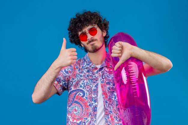 Unpleased young handsome curly man wearing sunglasses holding swim ring showing thumbs up and down on isolated blue wall