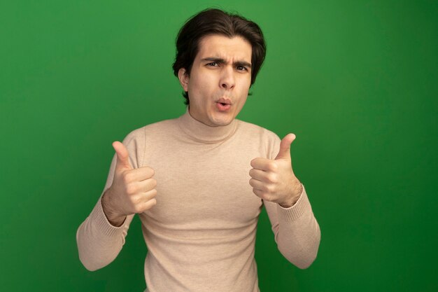 Unpleased young handsome guy showing thumbs up isolated on green wall