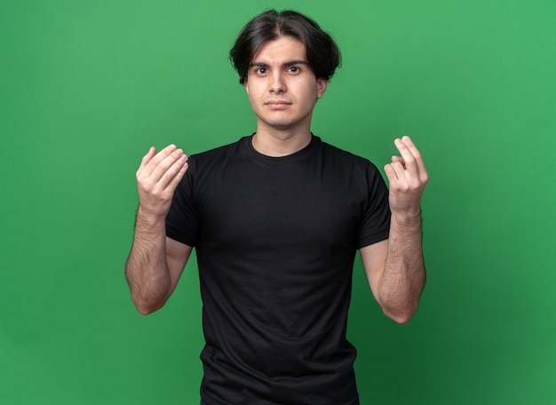 Unpleased young handsome guy wearing black t-shirt showing tip gesture isolated on green wall