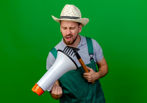 Free photo unpleased young handsome slavic gardener in uniform and hat holding speaker and rake looking at speaker isolated on green wall with copy space