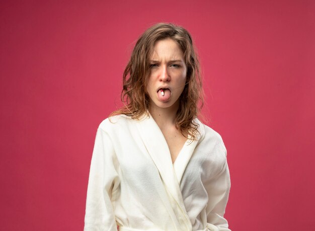 Unpleased young ill girl wearing white robe putting pill in mouth isolated on pink