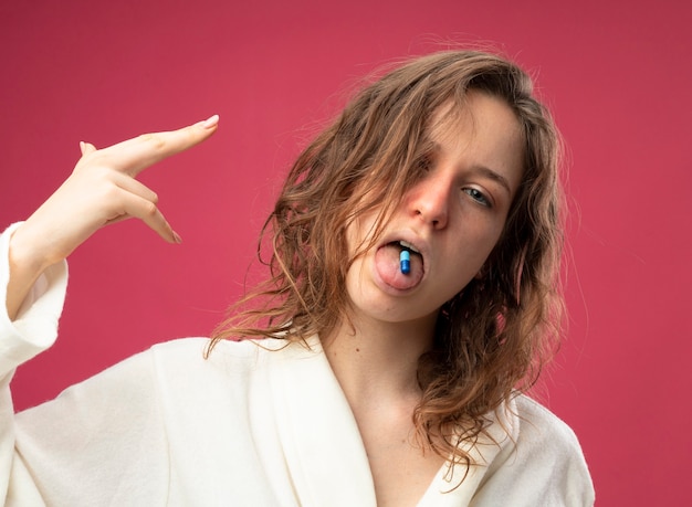 Unpleased young ill girl wearing white robe putting pills in mouth and showing suicide with pistol gesture isolated on pink