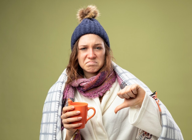 Free photo unpleased young ill girl wearing white robe and winter hat with scarf wrapped in plaid holding cup of tea showing thumb down isolated on olive green