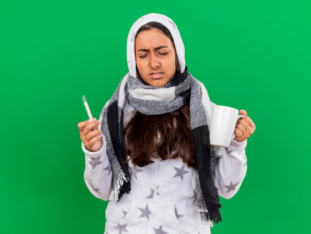 Free photo unpleased young ill girl with closed eyes putting on hood wearin scarf holding thermometer with cup of tea isolated on green background