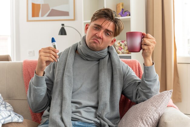 unpleased young ill man with scarf around neck holding thermometer and cup sitting on couch at living room