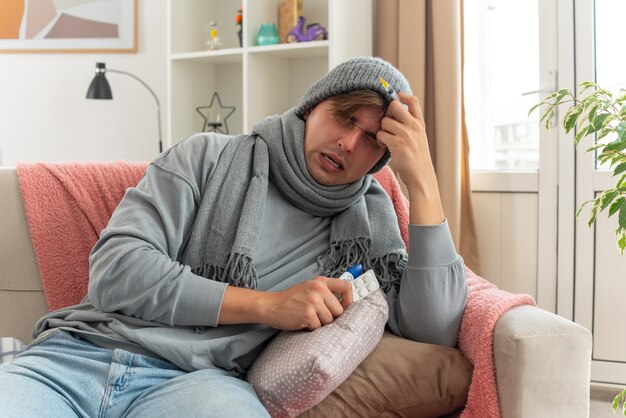 unpleased young ill man with scarf around neck wearing winter hat holding syringe and medicine blister pack and thermometer sitting on couch at living room