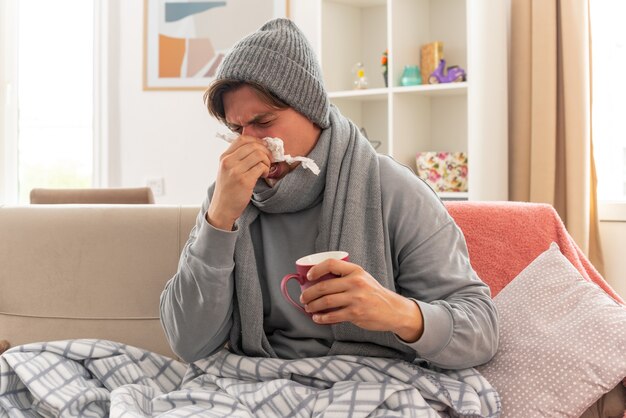 unpleased young ill man with scarf around neck wearing winter hat wipes his nose with tissue and holds cup sitting on couch at living room