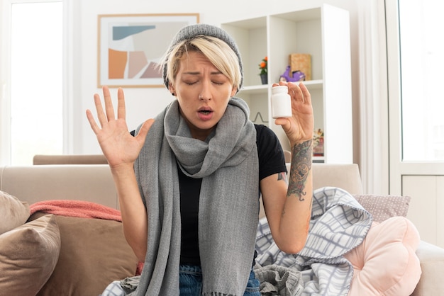 Free photo unpleased young ill slavic woman with scarf around her neck wearing winter hat holding medicine bottle and raising hand up sitting on couch at living room