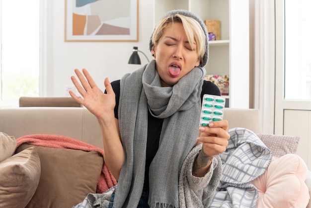 Free photo unpleased young ill slavic woman with scarf around her neck wearing winter hat stucks out tongue and holds medicine blister pack sitting on couch at living room