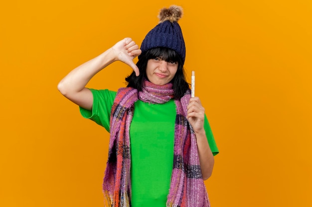 Unpleased young ill woman wearing winter hat and scarf holding thermometer looking at front showing thumb down isolated on orange wall with copy space