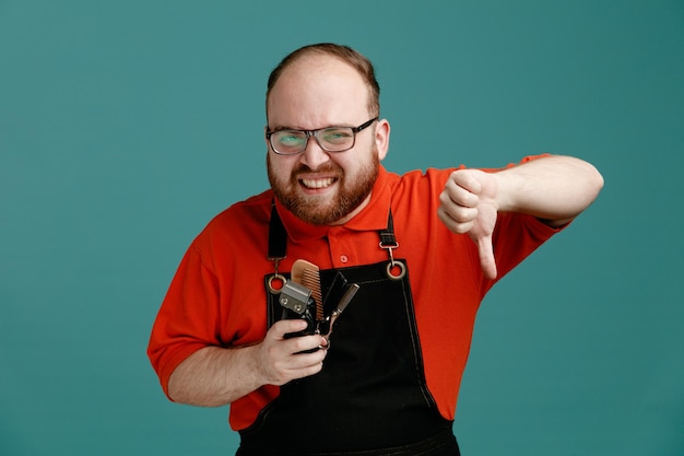 Free photo unpleased young male barber wearing glasses red shirt and barber apron holding barbering tools looking at camera showing thumb down isolated on blue background