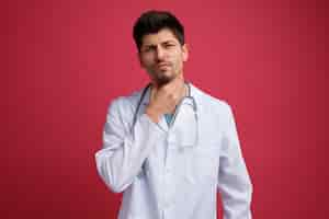 Free photo unpleased young male doctor wearing medical uniform and stethoscope around his neck keeping hand on neck looking at camera having sore throat isolated on red background