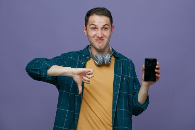 Unpleased young male student wearing headphones around neck looking at camera showing mobile phone and thumb down isolated on purple background