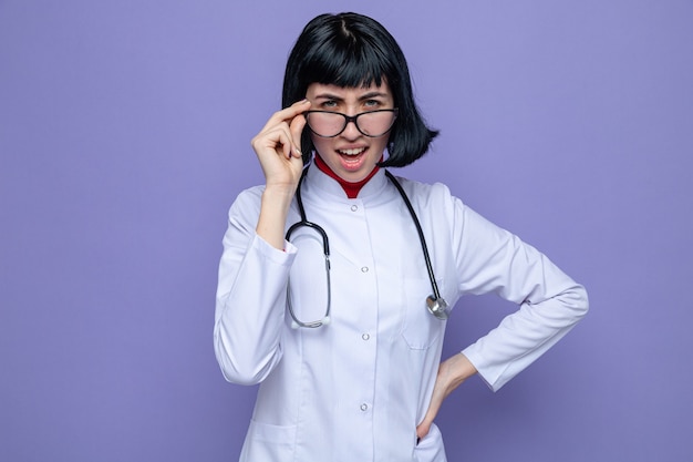 Unpleased young pretty caucasian girl with optical glasses wearing doctor uniform with stethoscope 