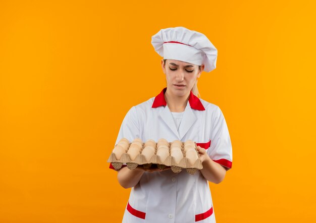 Unpleased young pretty cook in chef uniform holding and looking at carton of eggs isolated on orange space