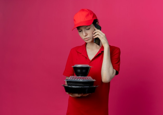 Free photo unpleased young pretty delivery girl wearing red uniform and cap talking on phone and holding and looking at food containers isolated on crimson background with copy space