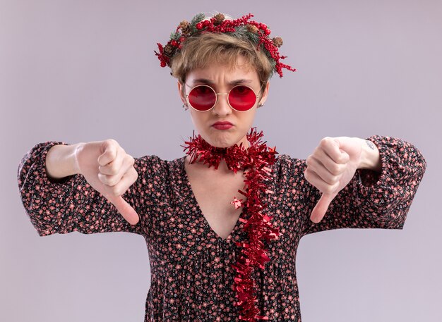 unpleased young pretty girl wearing christmas head wreath and tinsel garland around neck with glasses  showing thumbs down isolated on white wall