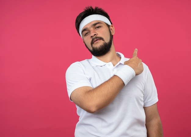 Unpleased young sporty man wearing headband and wristband points at behind isolated on pink wall with copy space